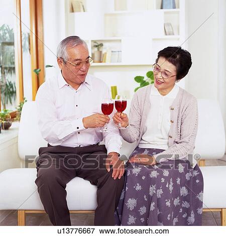 Stock Photo - Senior Couple Clinking Glasses of Red Wine, Sitting on a Sofa, Smiling, Front View. Fotosearch
