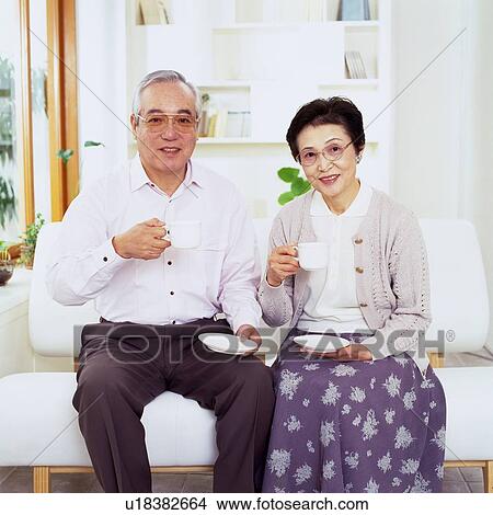 Picture - Senior Couple Sitting on the Sofa Having a Cup of Coffee, Looking at Camera, Smiling, Front View. Fotosearch
