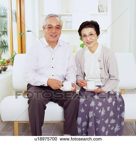 Stock Image - Senior Couple Sitting on the Sofa Having a Cup of Coffee, Looking at Camera, Smiling, Front View. Fotosearch