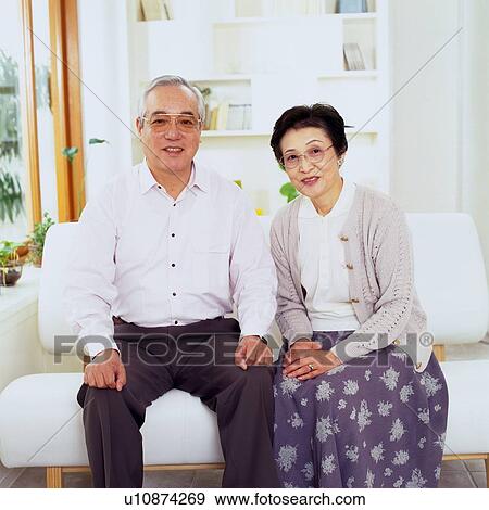 Stock Photo - Senior Couple Sitting on the Sofa Smiling, Looking at Camera, Front View. Fotosearch