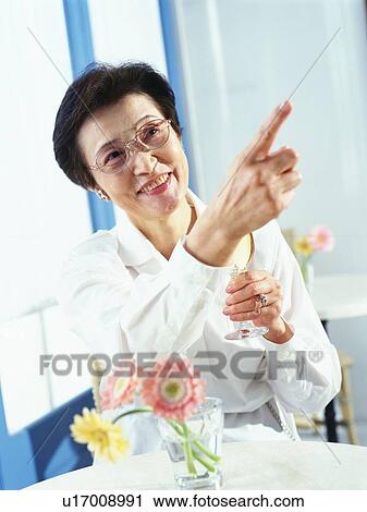 Stock Image - Senior woman holding a glass and pointing up, sitting by window, Smiling, Low Angle View. Fotosearch