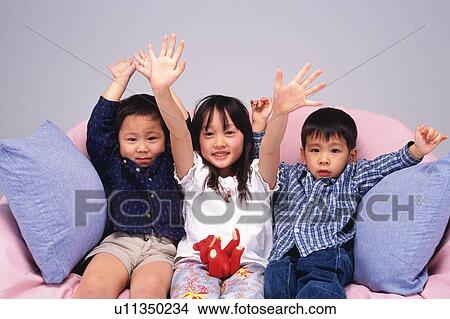 Picture -  Three Little Children, Sitting on a Sofa Waving Their Hands, Smiling, Looking at Camera, Front View. Fotosearch