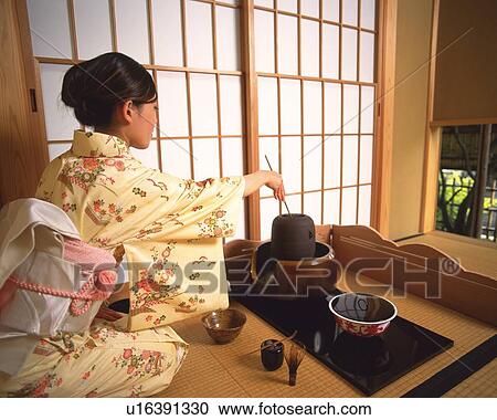 Stock Image - Woman in kimono scooping hot water from tea kettle with a bamboo dipper in a tea room, rear view, Japan. Fotosearch