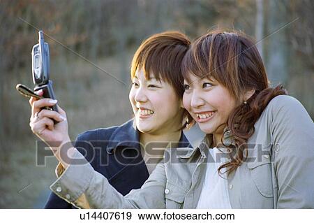 Stock Photo - Young women photographing themselves with mobile phone, smiling . Fotosearch