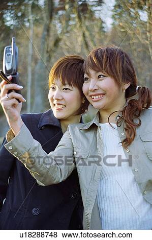Stock Photo - Young women photographing themselves with mobile phone, smiling . Fotosearch