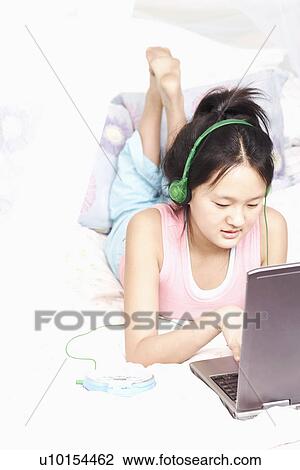 Stock Image - High angle view of a girl using a laptop listening to music on an MP3 player. Fotosearch