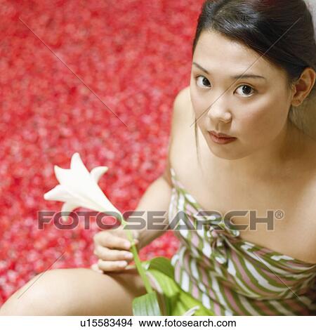Picture - High angle view of a young woman holding a flower and sitting near a hot tub filled with rose petals. Fotosearch