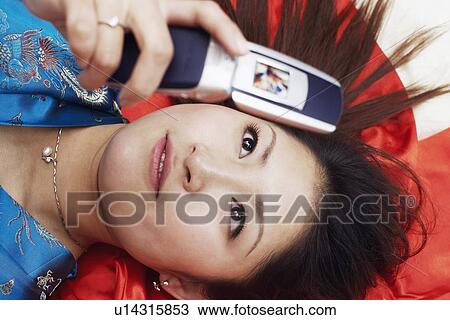 Stock Image - High angle view of a young woman lying on her back holding a mobile phone. Fotosearch