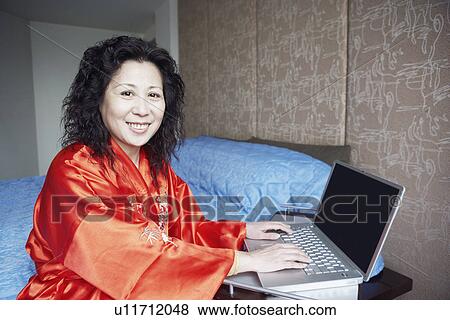 Stock Photo - Portrait of a mature woman using a laptop smiling in the bedroom. Fotosearch