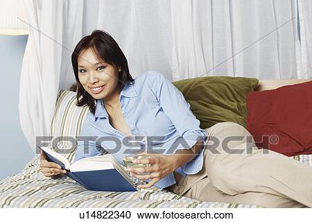 Stock Image - Portrait of a young woman holding a wineglass and a book on the bed. Fotosearch