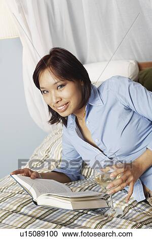 Stock Image - Portrait of a young woman holding a wineglass and a book on the bed. Fotosearch