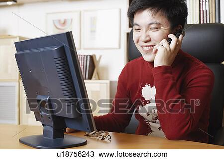 Stock Image - Close-up of a businessman sitting in front of a computer monitor using a mobile phone. Fotosearch