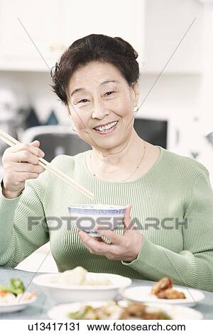 Stock Image - Portrait of a senior woman holding a pair of chopsticks and a bowl. Fotosearch
