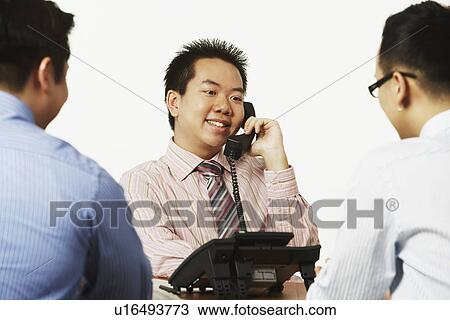 Stock Image - Close-up of a businessman talking on the telephone with two businessmen sitting in front of him. Fotosearch