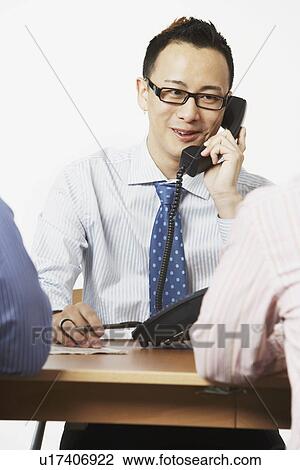 Stock Image - Close-up of a businessman talking on the telephone with two businessmen sitting in front of him. Fotosearch