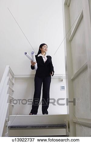 Stock Photo - Low angle view of a female architect standing on the staircase and holding rolled up blueprints. Fotosearch