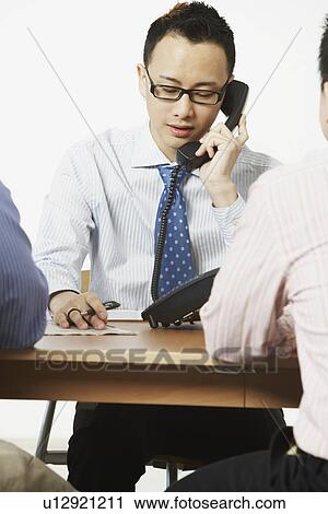 Close-up of a businessman talking on the telephone with two businessmen sitting in front of him View Large Photo Image Stock Image - Close-up of a businessman talking on the telephone with two businessmen sitting in front of him. Fotosearch