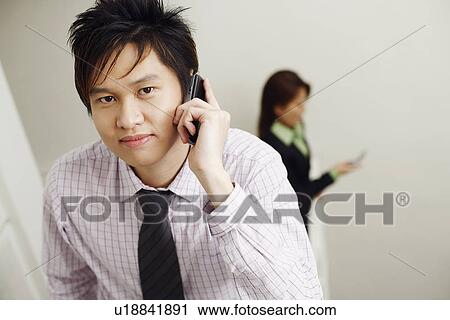 Stock Image - Portrait of a businessman talking on a mobile phone with a businesswoman in the background. Fotosearch