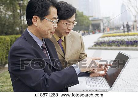 Stock Photo - Side profile of two businessmen using a laptop. Fotosearch