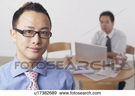 Stock Photo - Portrait of a businessman smiling with another businessman using a laptop behind him. Fotosearch