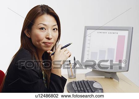 Stock Image - Portrait of a businesswoman sitting in front of a computer and holding a pen. Fotosearch