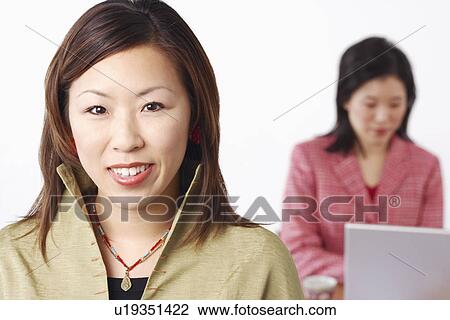 Stock Image - Portrait of a businesswoman smiling with another businesswoman using a laptop behind her. Fotosearch