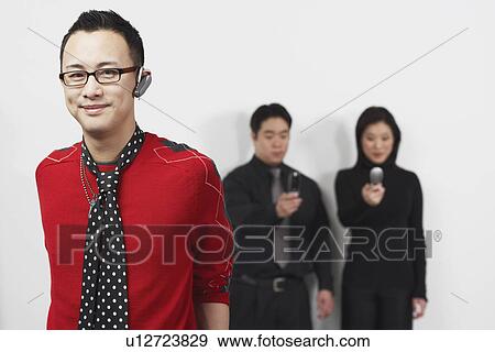 Stock Photo - Portrait of a young man using a hands free device with a businessman and businesswoman standing behind him. Fotosearch