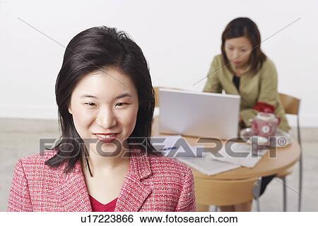 Stock Photograph - Portrait of a businesswoman smiling with another businesswoman using a laptop behind her. Fotosearch