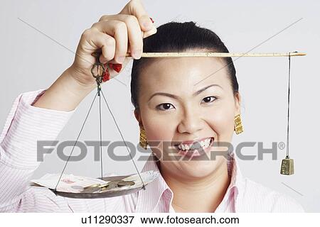 Stock Photo - Portrait of a businesswoman weighing paper currency with coins on a weighing scale and smiling. Fotosearch