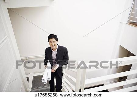 Stock Photo - Portrait of a businessman walking up stairs and carrying blueprints. Fotosearch