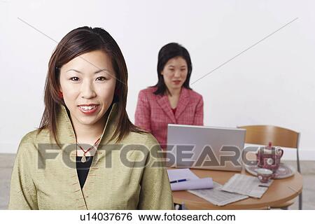 Stock Photograph - Portrait of a businesswoman smiling with another businesswoman using a laptop behind her. Fotosearch