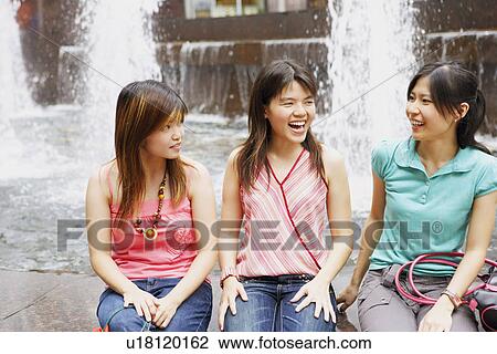 Stock Image - Three young women sitting together at the edge of a fountain and looking cheerful. Fotosearch