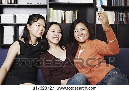 Stock Photo - Close-up of a mid adult woman sitting with two young women on a couch taking a photograph of themselves. Fotosearch