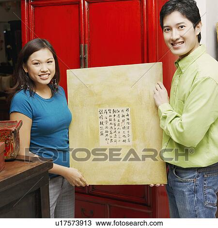 Stock Image - Portrait of a young couple holding a painting and smiling. Fotosearch