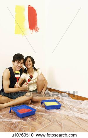 Stock Image - Portrait of a young couple sitting together on the floor and holding paintbrushes. Fotosearch