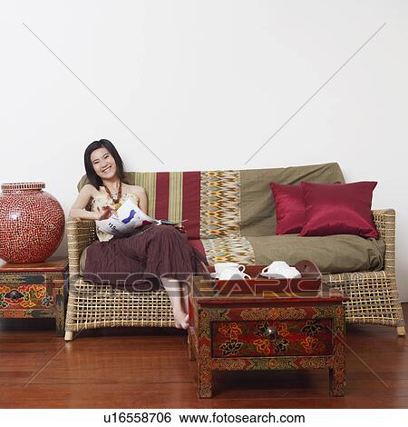Stock Photograph - Portrait of a young woman sitting on a couch and holding a magazine. Fotosearch