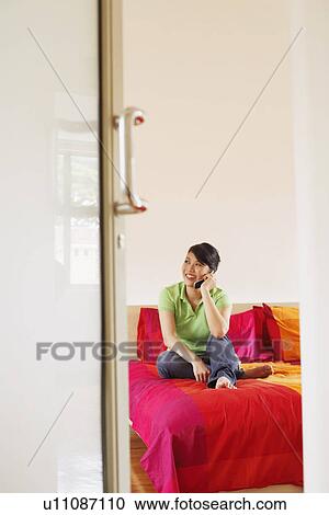 Stock Image - Young woman sitting on the bed and talking on a mobile phone. Fotosearch
