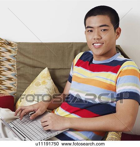 Stock Image - Portrait of a young man sitting on a couch and using a laptop. Fotosearch