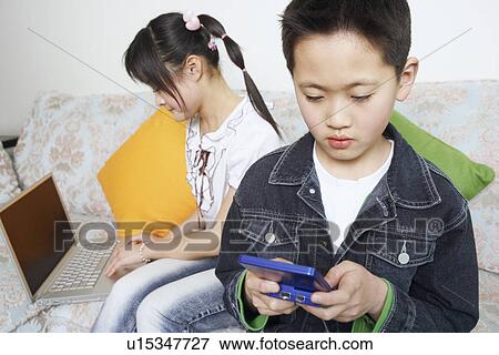 Boy playing a video game beside his sister using a laptop View Large Photo Image Stock Photo - Boy playing a video game beside his sister using a laptop. Fotosearch