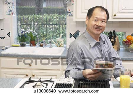Stock Image - Portrait of a senior man holding a newspaper in the kitchen. Fotosearch