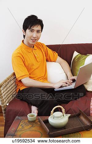 Portrait of a young man sitting on a couch and using a laptop View Large Photo Image Picture - Portrait of a young man sitting on a couch and using a laptop. Fotosearch