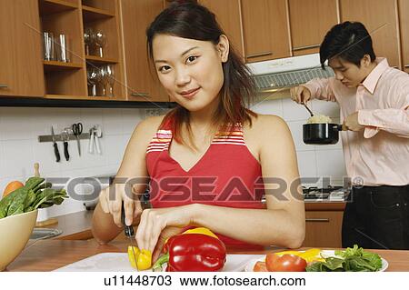Stock Image - Portrait of a young woman cutting vegetables in the kitchen with a young man eating noodles in the background. Fotosearch