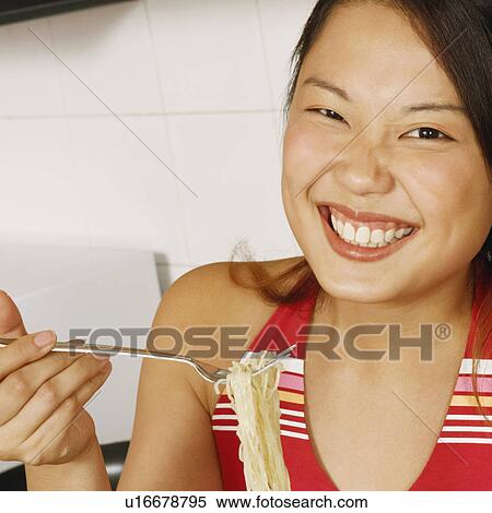 Stock Photography - Portrait of a young woman holding noodles with a fork at a kitchen counter. Fotosearch
