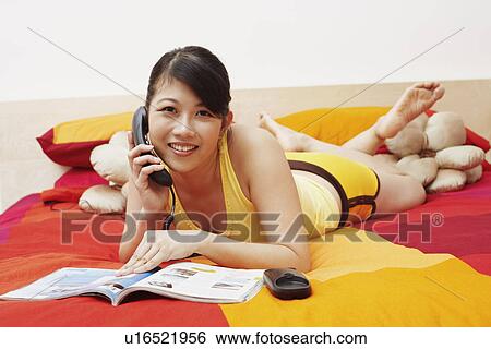 Stock Photograph - Portrait of a young woman talking on the telephone and lying on the bed. Fotosearch