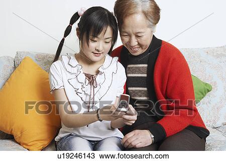 Stock Image - Young woman sitting together with her grandmother holding a mobile phone. Fotosearch