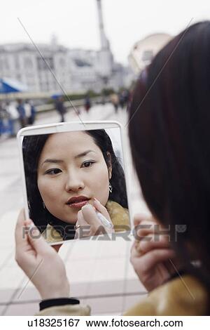 Stock Photo - Close-up of a young woman looking into a mirror applying lipstick. Fotosearch