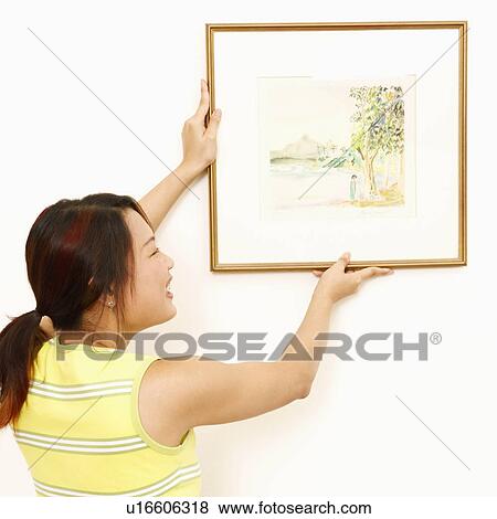 Stock Photo - Rear view of a young woman mounting a picture frame on the wall and smiling. Fotosearch