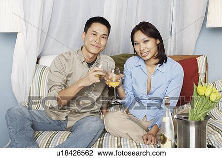 Stock Image - Portrait of a young couple sitting on the bed toasting with wineglasses. Fotosearch