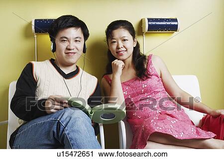 Stock Image - Portrait of a young man and a mid adult woman sitting on chairs listening to music. Fotosearch