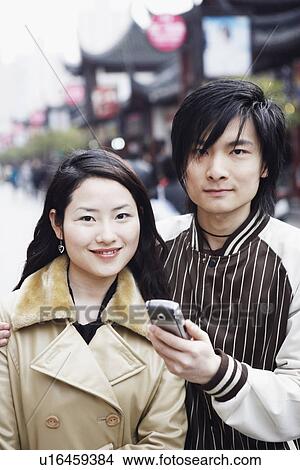 Picture - Portrait of a young man holding a mobile phone with a young woman smiling. Fotosearch